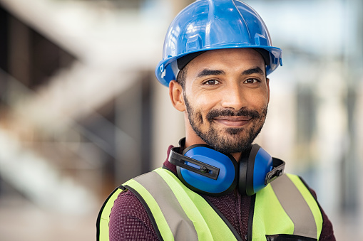 Portrait of satisfied construction site manager wearing safety vest and blue helmet at construction site. Young middle eastern architect watching construction site with confidence looking at camera. Indian manual worker on a industrial building with copy space.
